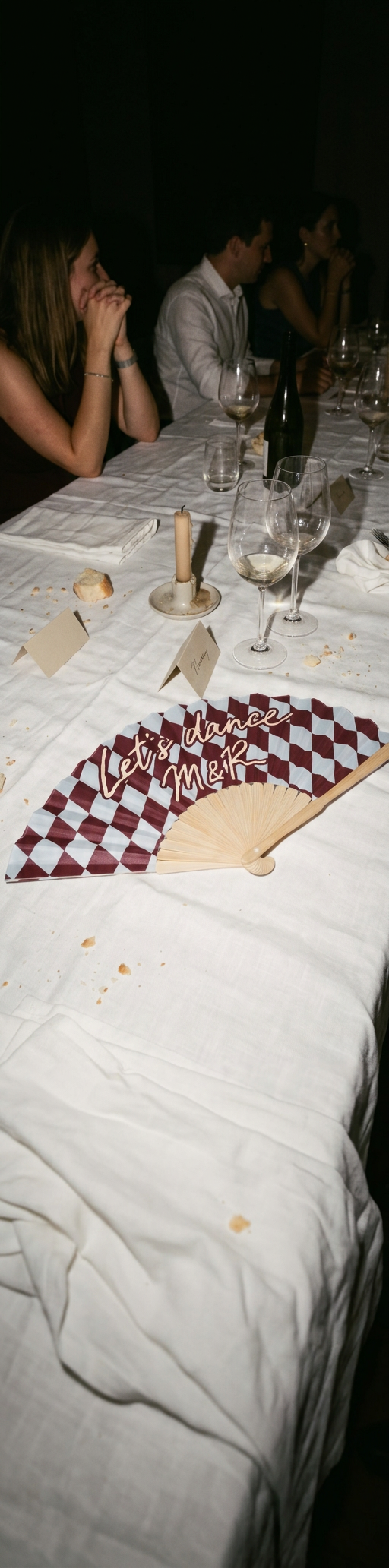 A burgundy and pale blue harlequin hand fan lettered 'Let's dance M&R' resting on a set banquet table with wine glasses and place cards
