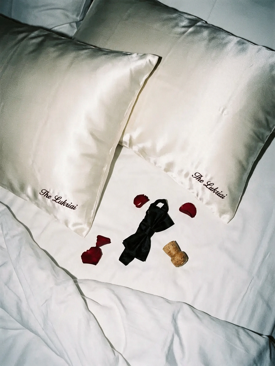 Two ivory satin pillowcases embroidered ‘The Lukriai’ in burgundy script, resting on a white bed