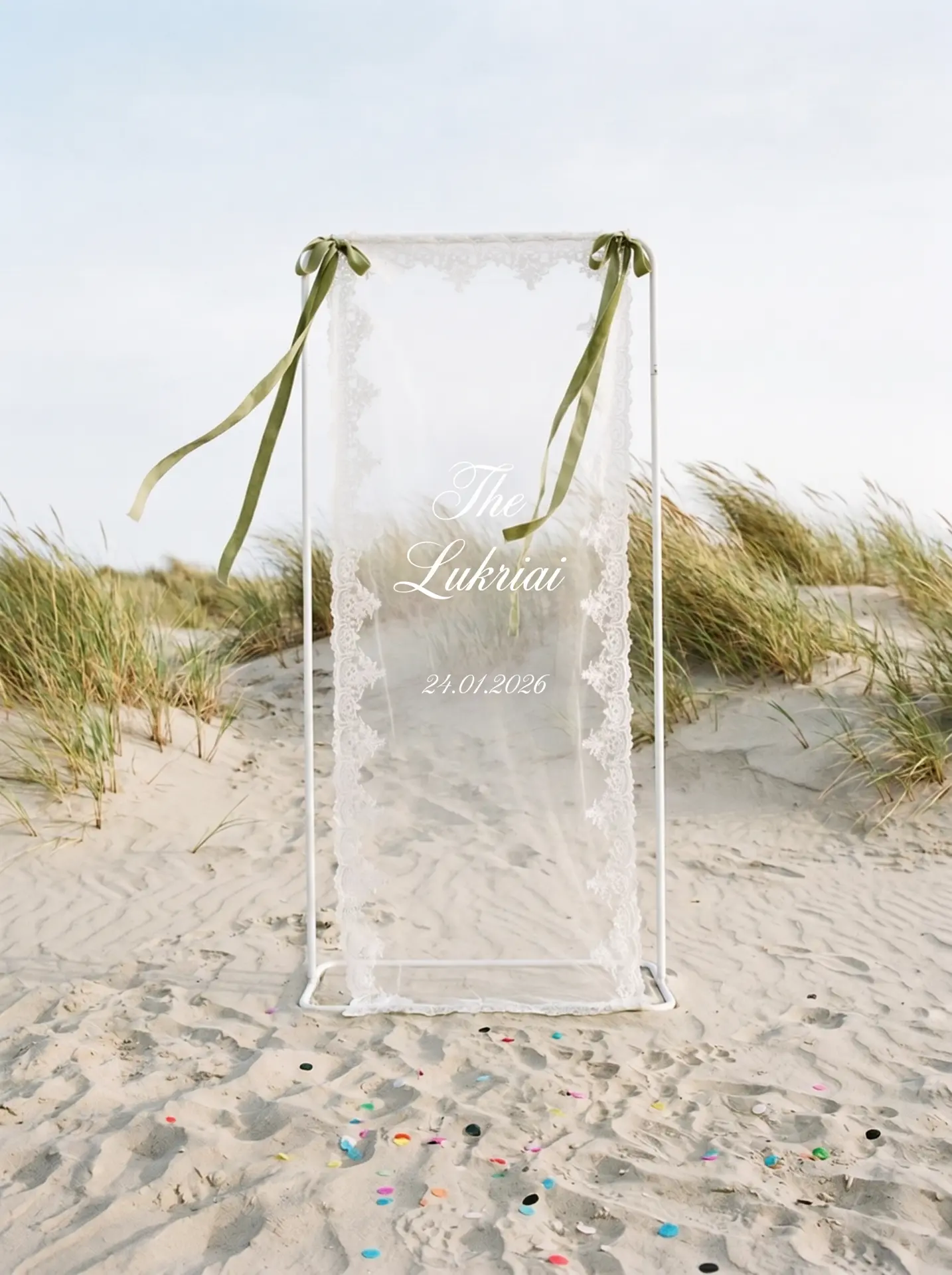 A lace-edged tulle welcome sign printed The Lukriai 24.01.2026 freestanding on a white metal stand among beach dunes with confetti on the sand